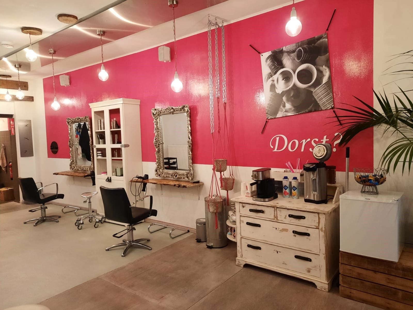 De salon van Haar&Mel: een felroze muur met zilveren barokspiegels, zwarte kappersstoelen, hanglampjes en een koffiehoek op een witte commode